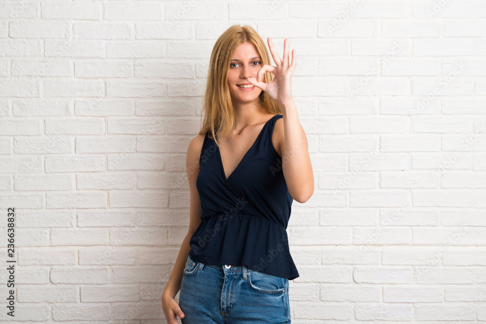 Blonde woman showing an ok sign with fingers. Face of happiness and satisfaction on white brick wall background Blonde woman showing an ok sign with fingers. Face of happiness and satisfaction on white brick wall background