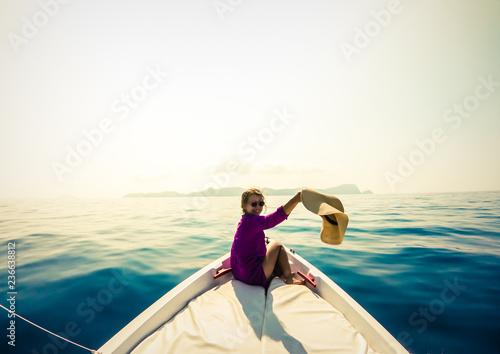 Young woman sitting on bow of boat in the middle of the sea. Happy girl with hat raising hands enjoying freedom travel.