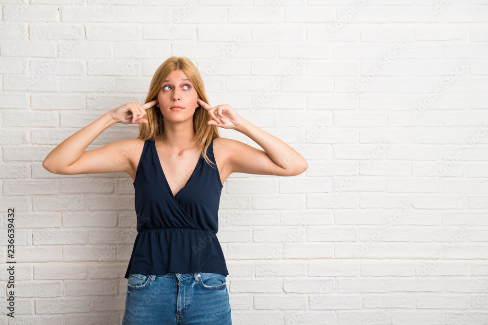 Blonde woman covering both ears with hands on white brick wall background Blonde woman covering both ears with hands on white brick wall background