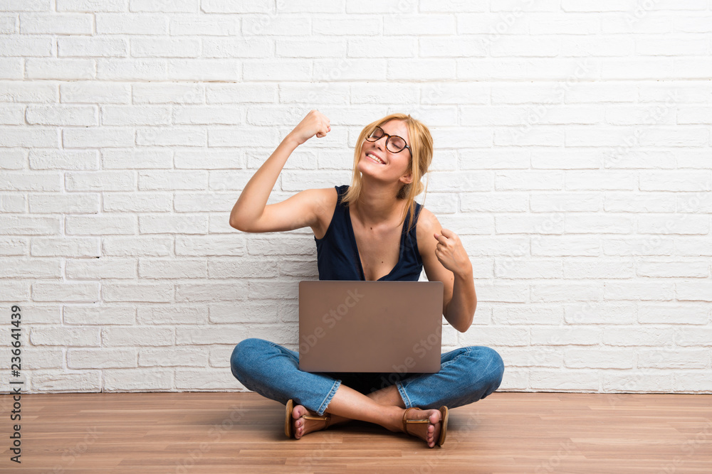 Blonde girl sitting on the floor with her laptop celebrating a victory on white brick wall background Blonde girl sitting on the floor with her laptop celebrating a victory on white brick wall background