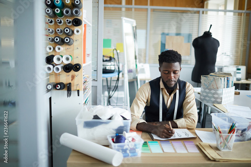 Young serious designer of clothes sitting by desk in his workshop and sketching new fashion models photo