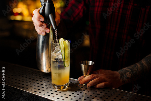 Barman pouring soda into a glass with cucumber making fresh cocktail