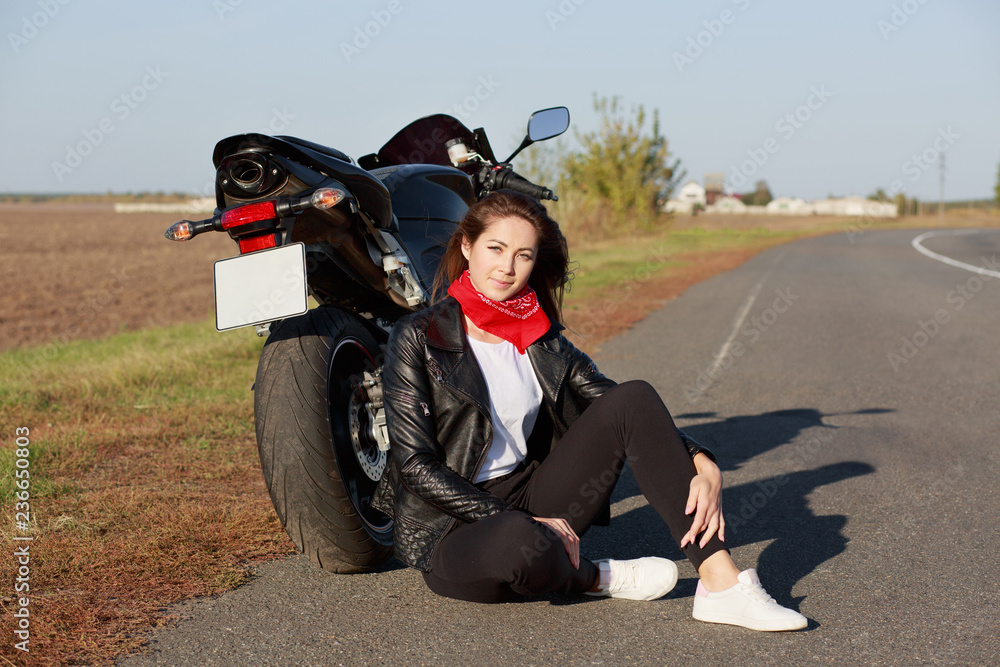 Naklejka premium Relaxed carefree female motorcyclist wears black clothes and white trainers, sits at asphalt near motorbike, enjoys adventure and racing in countryside, prepares for motorcross. Leisure concept