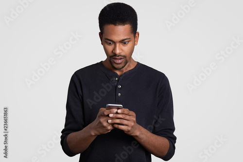Astonished black man with curly hair, holds mobile phone, recieves text message, feels surprised and amazed, wears casual clothes, isolated over white background. People, reaction and technology © sementsova321