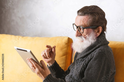 Senior bearded man in glasses wearing grey pullover sitting on a yellow sofa in his light living room and swipping facebook news on the digital tablet.
