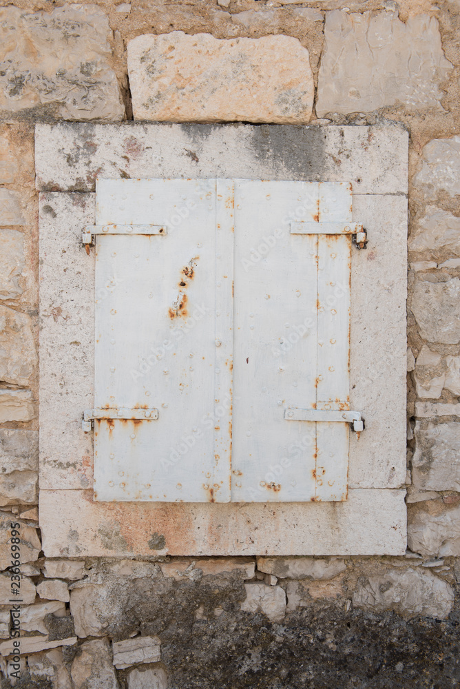 Naklejka premium Very old closed window with white wooden shutters. Window with stone frame on a stone house.