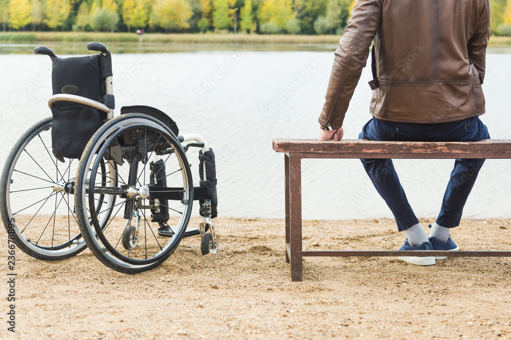 Obraz premium A young man sits on a bench by the lake, next to his wheelchair.