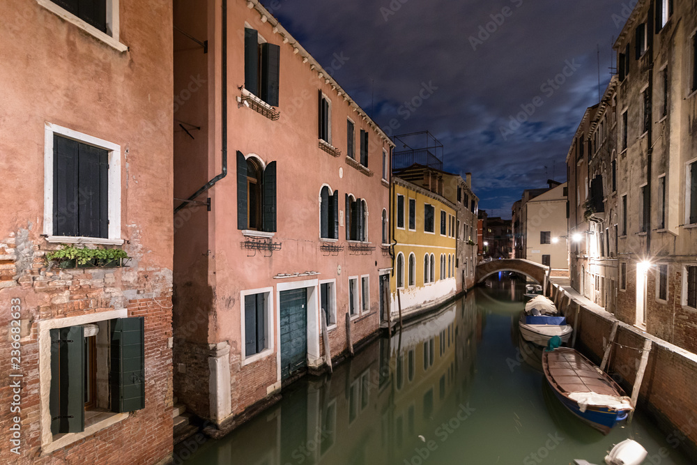 A narrow canal - street with boats in Venice at night, Italy.