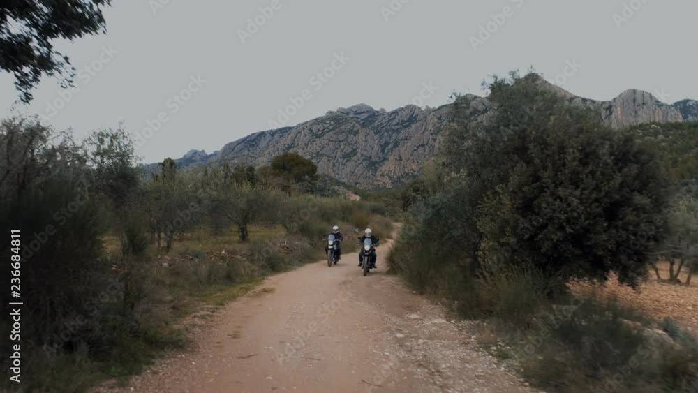 Two motorcycles drive towards camera on quiet empty countryside gravel road. Mountains in background in beautiful scenery during offroad enduro or motocross adventure. Extreme sports Two motorcycles drive towards camera on quiet empty countryside gravel road. Mountains in background in beautiful scenery during offroad enduro or motocross adventure. Extreme sports