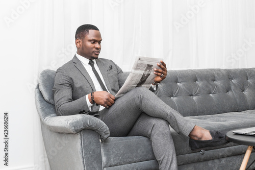 Fototapeta Naklejka Na Ścianę i Meble -  Young African American businessman in a gray suit reading a newspaper while sitting on a sofa.