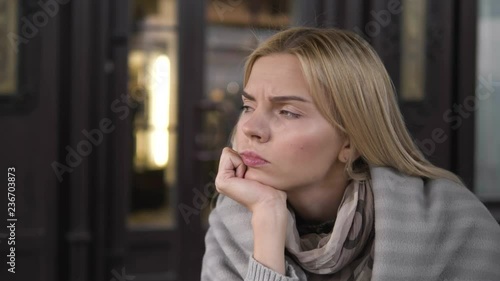 A young beautiful blonde trendy dressed woman thinking about something resting her chin on her arm sitting in a cozy street cafe.
