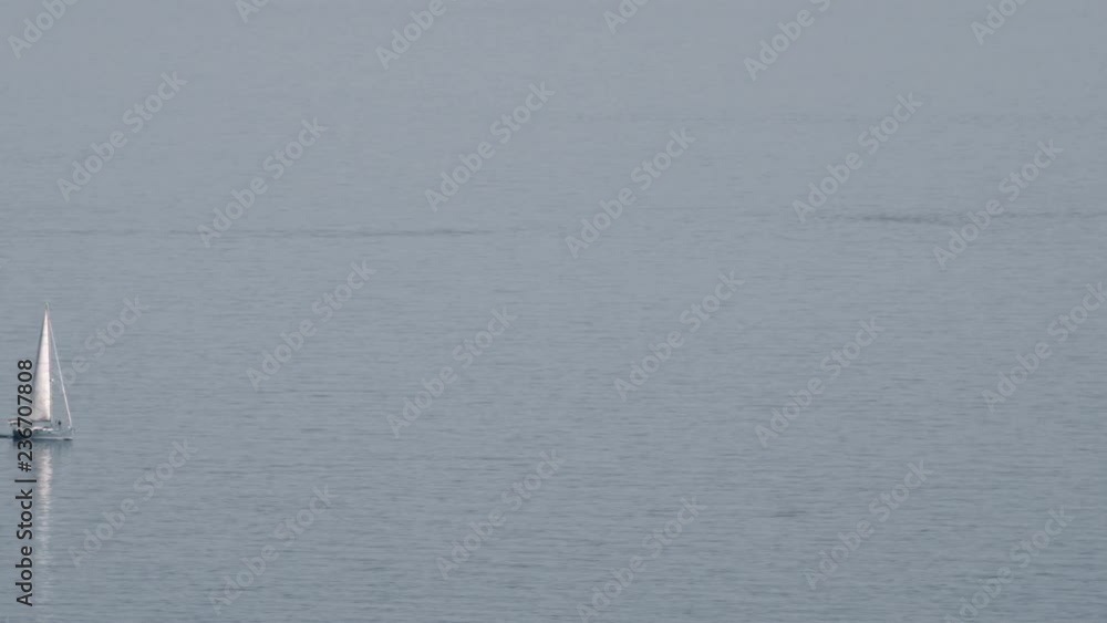 A lonely white yacht is slowly floating on the quite sea surface. The day is clear, there is no wind so nothing is bothering the water and the white sail A lonely white yacht is slowly floating on the quite sea surface. The day is clear, there is no wind so nothing is bothering the water and the white sail