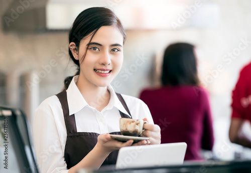 Young beautiful barista wearing brown apron holding hot coffee cup served to customer with smiling face at bar counter,Cafe restaurant service concept