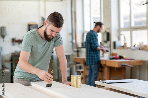 young worker in a carpenter's workshop painting wood with brush