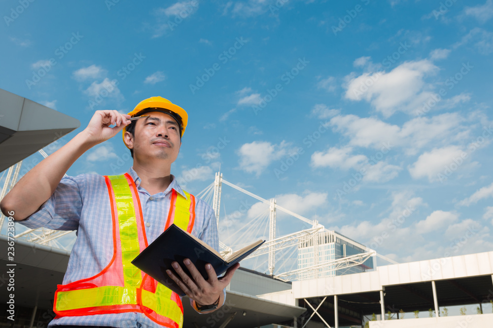 Young engineer wearing yellow helmet safety hold a work book look up to sky in front of construction building. Young engineer wearing yellow helmet safety hold a work book look up to sky in front of construction building.