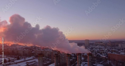 A large boiler room is located in the middle of a snowy city. From her five huge pipes, smoke and steam are pulled out of the air. It all sneaks up with a red sunshine.