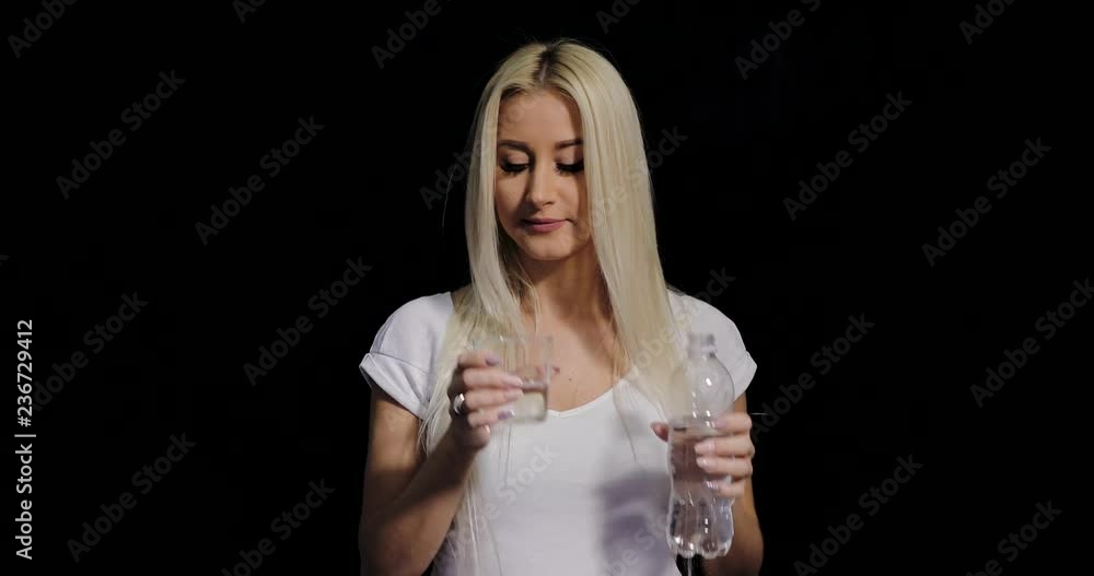 healthy blond woman pouring fresh water from a bottle into a glass and smiling on camera, over black background healthy blond woman pouring fresh water from a bottle into a glass and smiling on camera, over black background