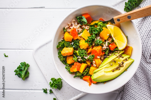 Vegan salad with rice, kale, baked pumpkin, carrots and avocado in white bowl on a white wooden background.