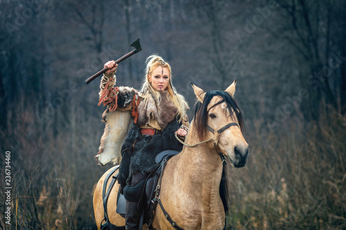 Fotografie Scandinavian viking woman warrior riding horse holding ax above head looking threateningly