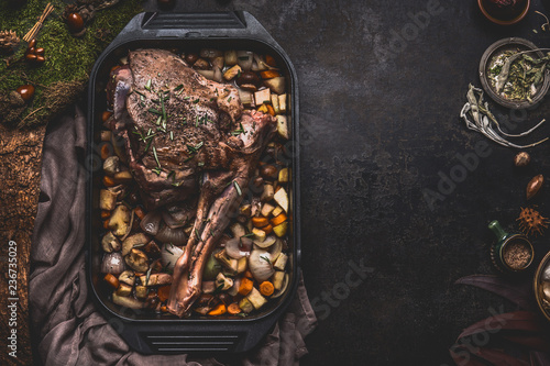 Cooking preparation of venison roast. Leg of deer with bone in cast iron pan with gut vegetables on dark kitchen table background with herbs and spices. Copy space for your recipe or design