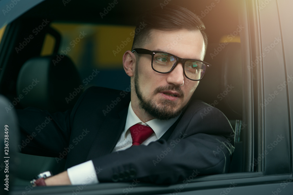 Portrait of successful businessman sitting at the wheel of an expensive car. Young bearded man wearing formal attire and stylish glasses driving a car