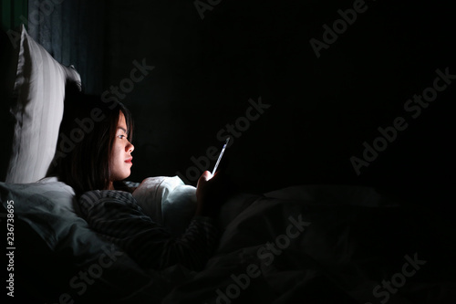 Asian woman using smartphone for checking social media with icon or hologram at night on the bed in dark room, Using smartphone in dark may be causes of eye pain symptom