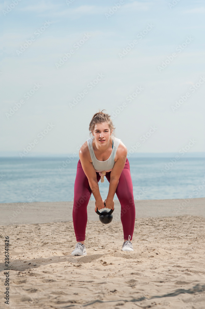 Young attractive woman dressed in pink leggings doing training trying hard in exercises with a kettle bell by the seaside. Fitness woman working out at the beach. Fitness Concept. Young attractive woman dressed in pink leggings doing training trying hard in exercises with a kettle bell by the seaside. Fitness woman working out at the beach. Fitness Concept.