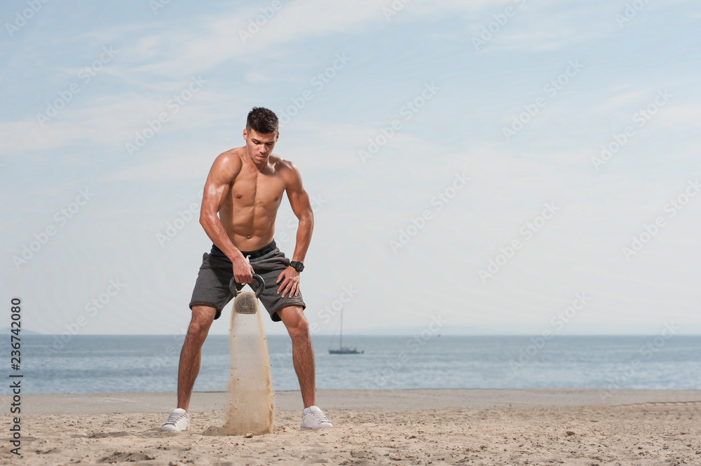 High intensive fitness workout. Young muscular man training with kettle bell on the beach against blue sky.