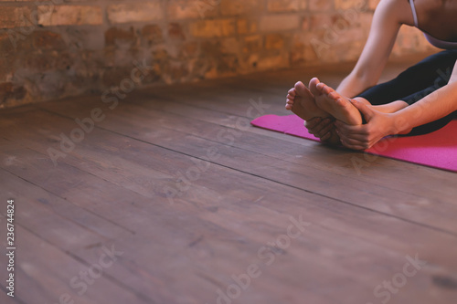 Close-up of a girl doing an asana bending forward with her hands towards her legs. Sitting on the floor on a  pink yoga mat.