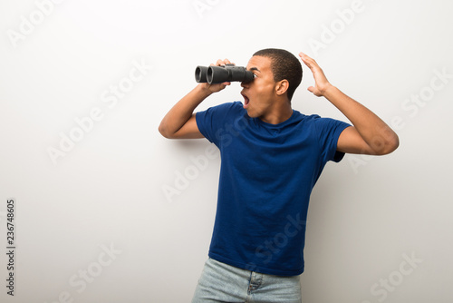 Young african american man on white wall and looking in the distance with binoculars © luismolinero