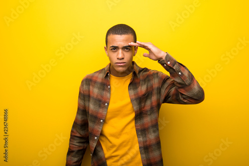 Young african american man on vibrant yellow background looking far away with hand to look something