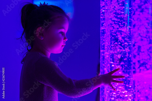 Child in therapy sensory stimulating room, snoezelen. Child interacting with colored lights bubble tube lamp during therapy session. photo