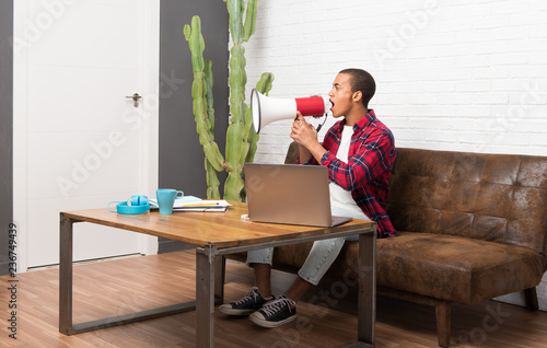 African american man with laptop in the living room shouting through a megaphone to announce something in lateral position