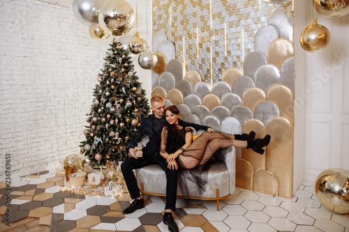 Christmas party concept. Stylishly dressed man and woman exchange gifts in a beautiful gold interior of the living room against the Christmas tree.