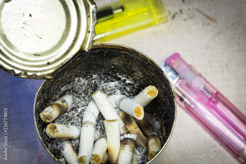 Cigarette tobacco butts and ashes in a tin can with pink and yellow lighters in the background. 