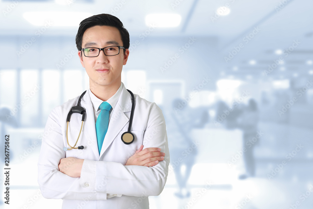Doctor posing with arms crossed, medical staff working on blurry background.