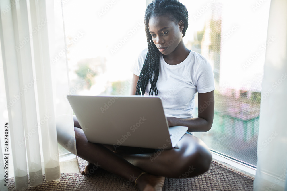 Beautiful young Afro American girl is working with a laptop and smiling while sitting on window sill at home Beautiful young Afro American girl is working with a laptop and smiling while sitting on window sill at home