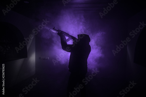 Fototapeta Naklejka Na Ścianę i Meble -  Silhouette of man with assault rifle ready to attack on dark toned foggy background or dangerous bandit in black wearing balaclava and holding gun in hand. Shooting terrorist with weapon theme decor