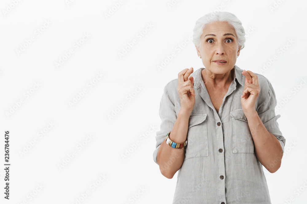 Waist-up shot of energzied worried hopeful granny with white hair in casual shirt clenching teeth crossing fingers for good luck watching lottery on tv and hoping win money posing over grey background Waist-up shot of energzied worried hopeful granny with white hair in casual shirt clenching teeth crossing fingers for good luck watching lottery on tv and hoping win money posing over grey background