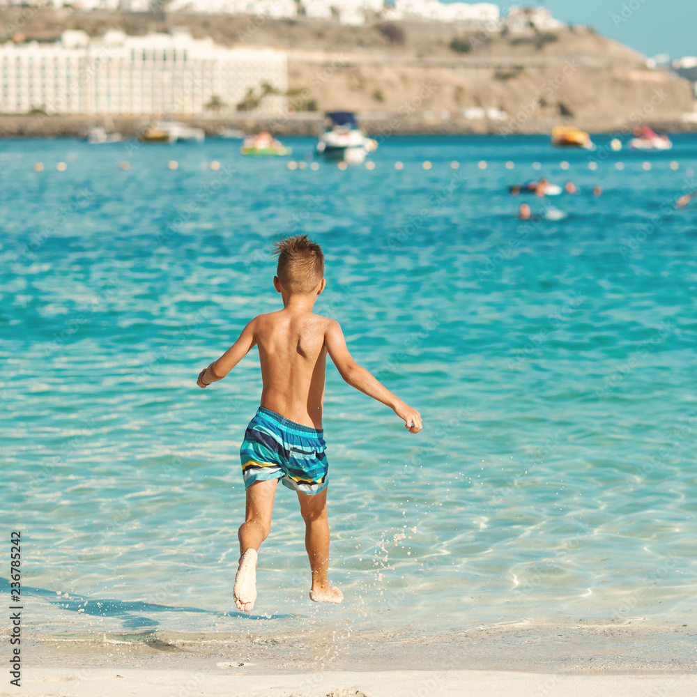 Active European boy in a striped swimming shorts is running towards the sea. He is having fun spending his holidays on the seashore. Active European boy in a striped swimming shorts is running towards the sea. He is having fun spending his holidays on the seashore.