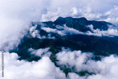 view from above, from the peak or from an airplane, to a wooded mountain valley with settlements, partly hidden by cumulus clouds
