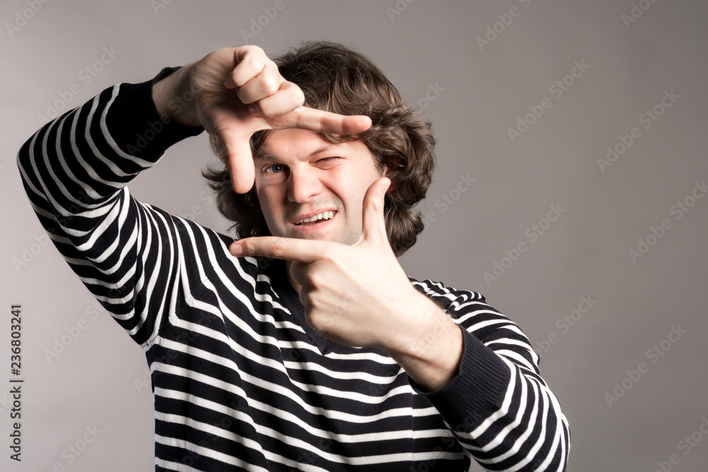 Young handsome dark curly hair man focusing with fingers, framing with hands looking at camera and gesturing finger frame isolated on grey background. Close up portrait. Studio shot. Young handsome dark curly hair man focusing with fingers, framing with hands looking at camera and gesturing finger frame isolated on grey background. Close up portrait. Studio shot.