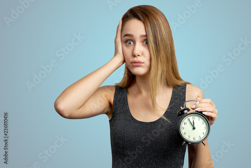 The girl holds her head, sighs, realizes that she was late. Clock at five minutes to twelve. Isolated on blue background in Studio