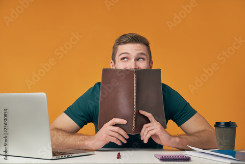 Happy european male student, has positive expression, holds brown book. Man rejoices successfully passed learned subject, isolated over orange background