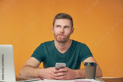 an attractive man in a t-shirt with a smartphone in his hands sitting at his Desk thinking and looking up. Isolated Studio portrait