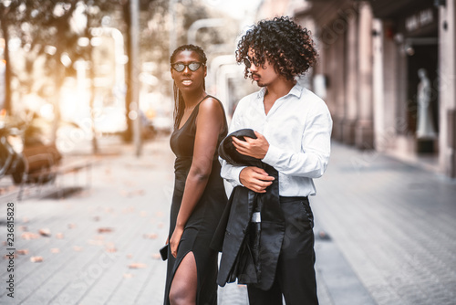 An interracial couple on the sidewalk: an African girl with braids in a black dress and a curly Asian guy with curly hair, in fancy sunglasses and with the blazer from his formal suit in hands photo