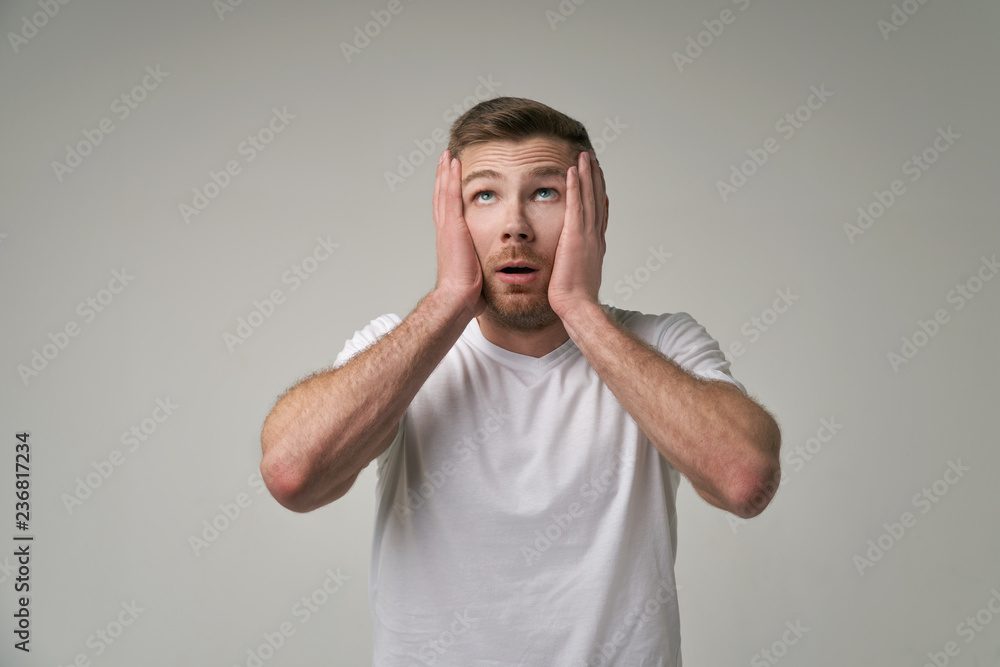 The astonished man looks up, isolated over white concrete wall. Bearded young man with blue eyes notices something unexpected upwards, keeps mouth opened. Shock concept The astonished man looks up, isolated over white concrete wall. Bearded young man with blue eyes notices something unexpected upwards, keeps mouth opened. Shock concept