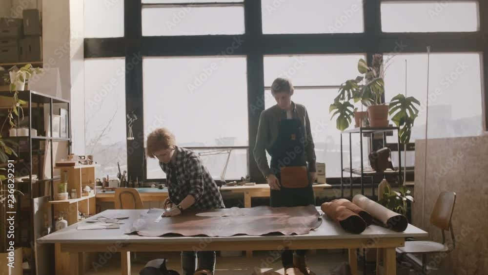 Man in apron walking into manufacturing workshop where craftswoman is cutting leather on a table to make handmade goods. Man in apron walking into manufacturing workshop where craftswoman is cutting leather on a table to make handmade goods.