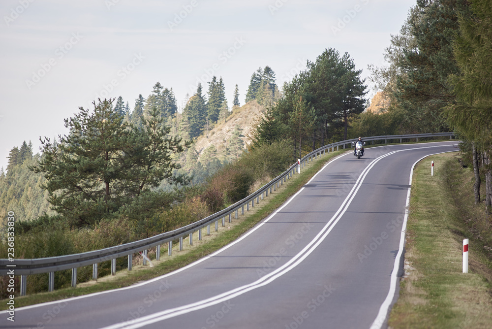 Fototapeta premium Alone motorcyclist in black leather outfit riding cruiser motorbike along twisted road on background foggy distant green woody hills under morning sky.