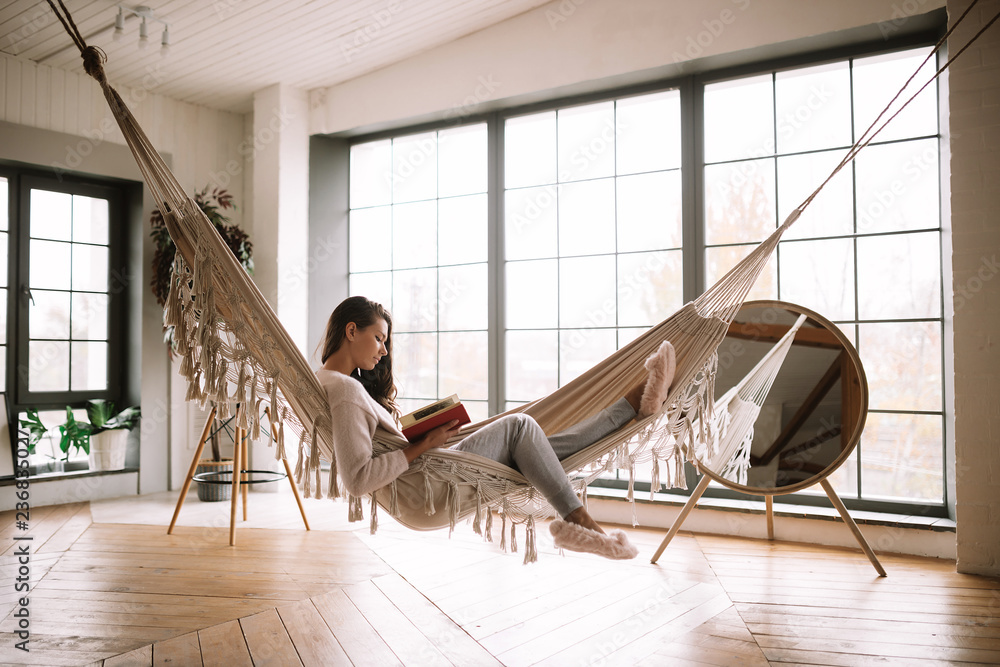 Dark-haired girl dressed in pants, sweater and warm slippers reads a book lying in a hammock in a cozy room with wooden floor and panoramic windows and a round mirror on the floor - obrazy, fototapety, plakaty 